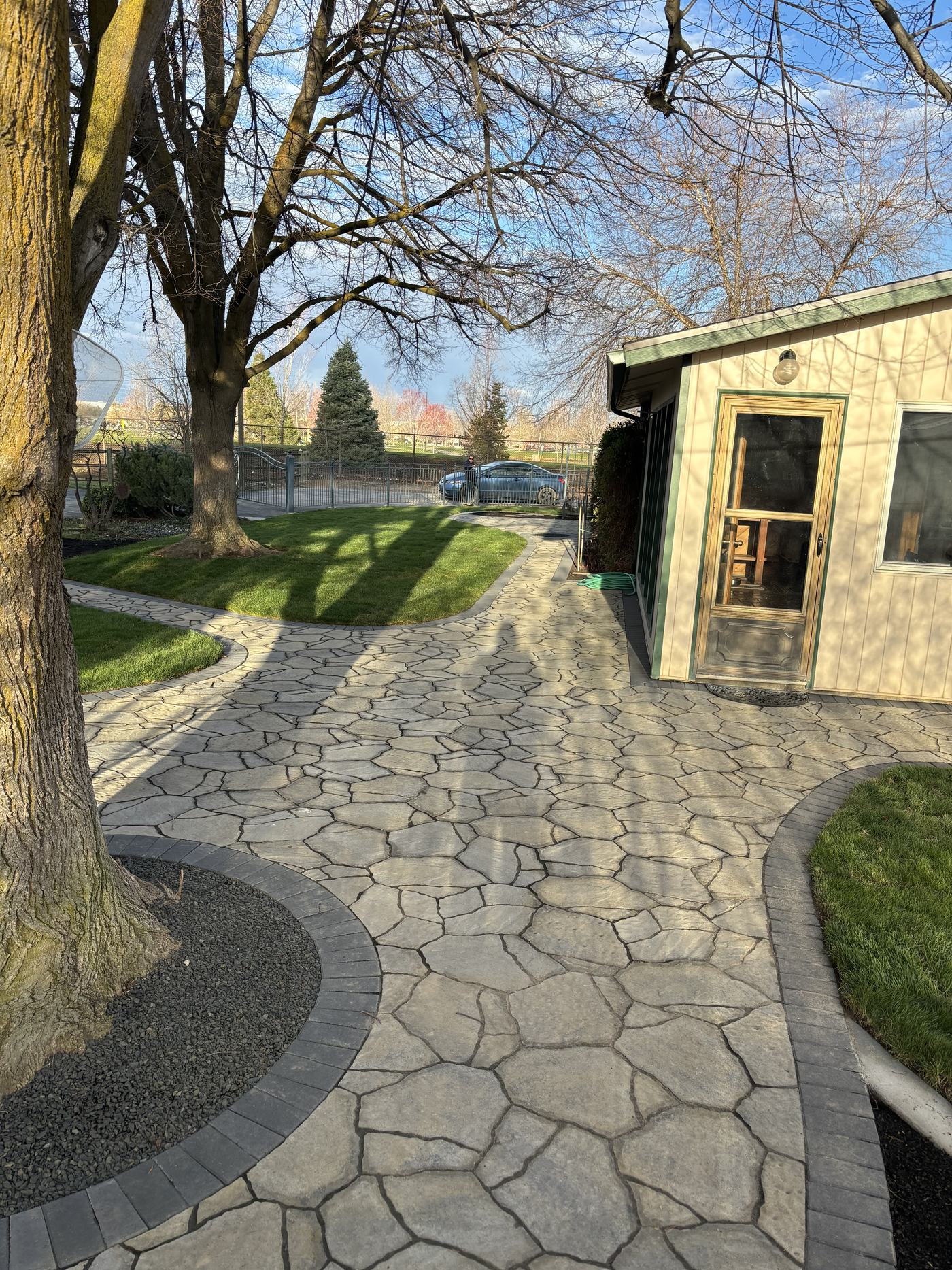 An irregular flagstone patio and path under mature shade trees, curving toward a timber outbuilding.