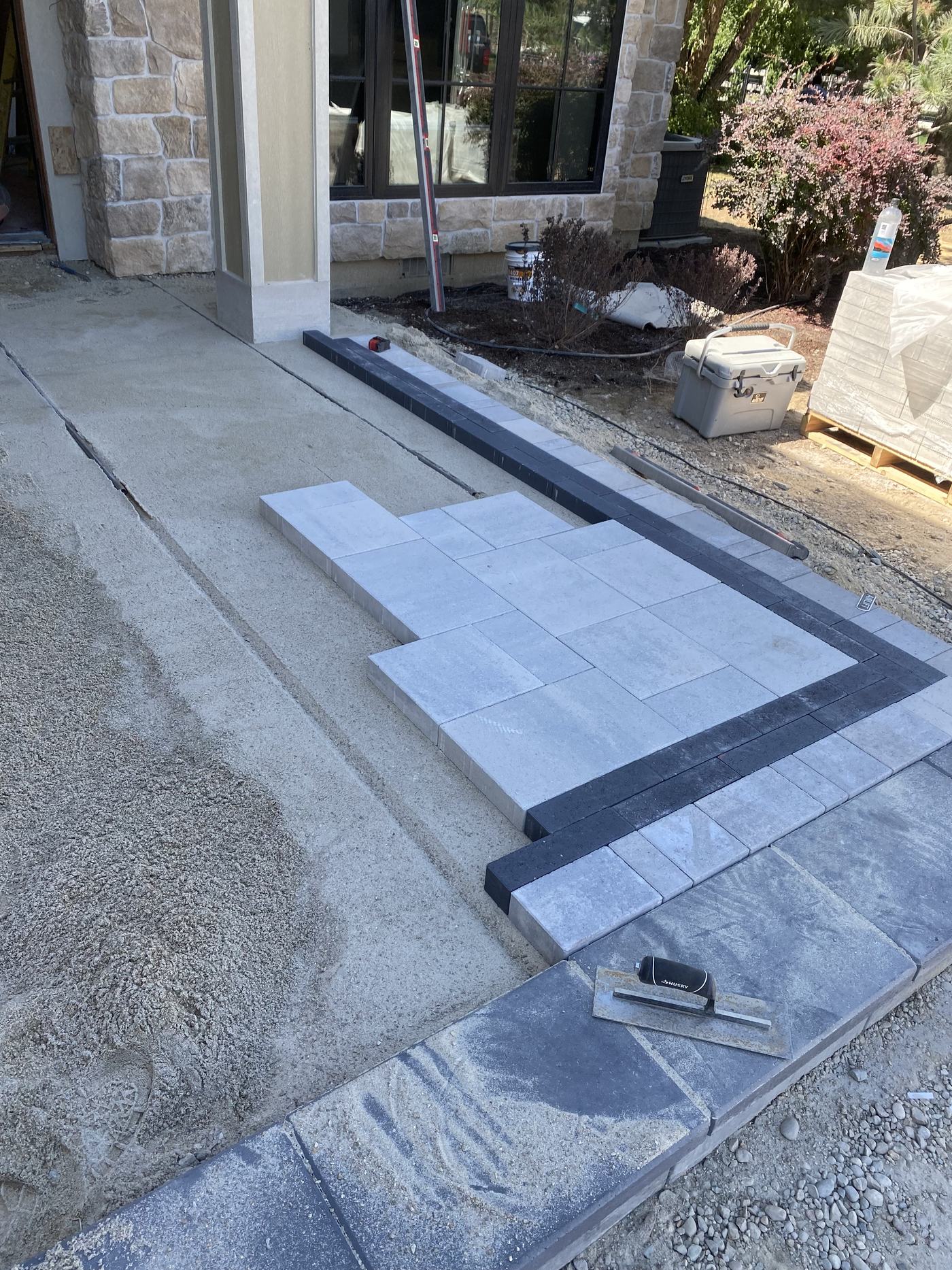 Large-format pavers being set into a screeded bed against a dark border course, on a patio under construction.