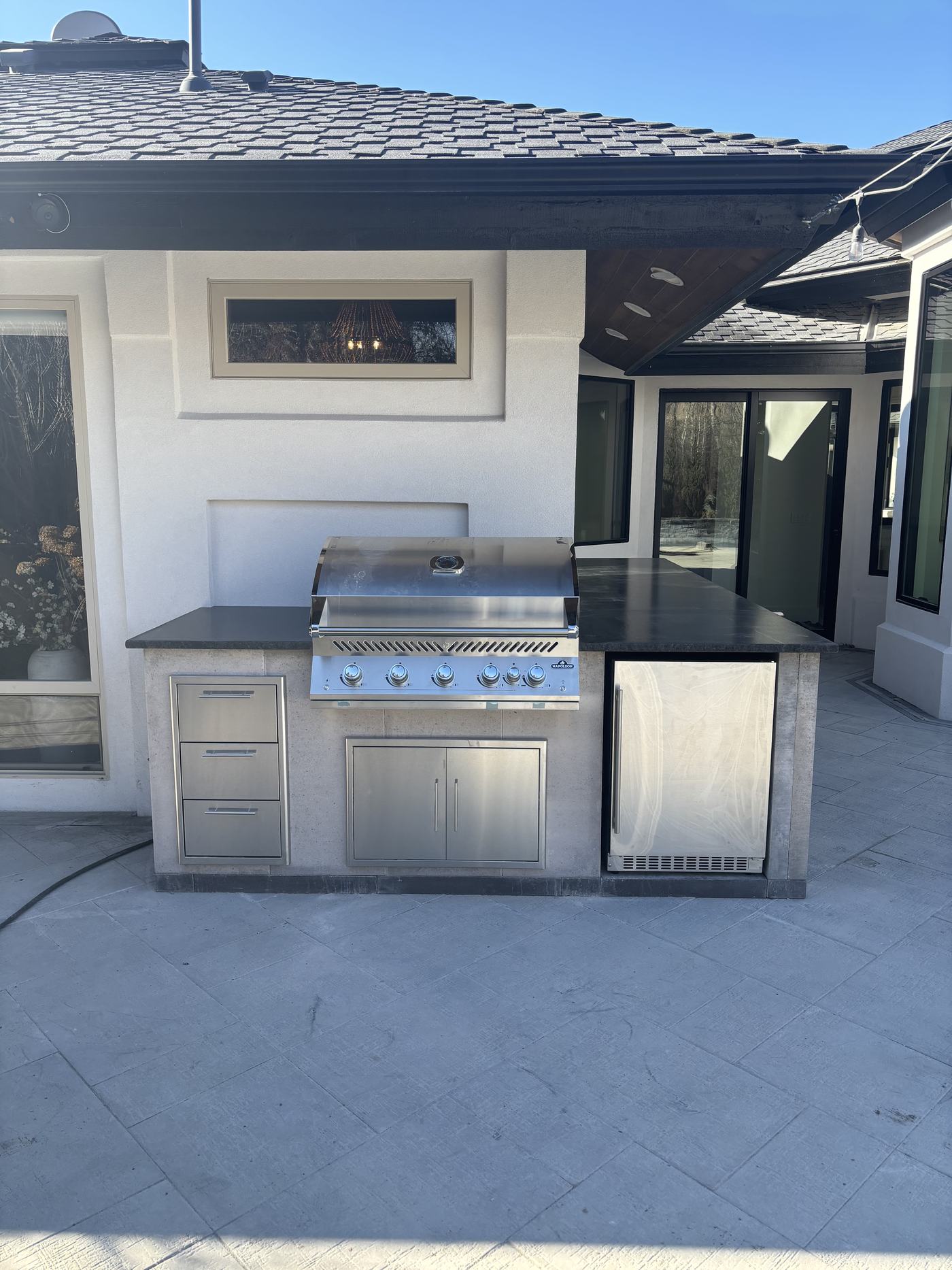 A built-in outdoor kitchen with a stainless gas grill, dark stone countertop and under-counter refrigerator under a covered patio.