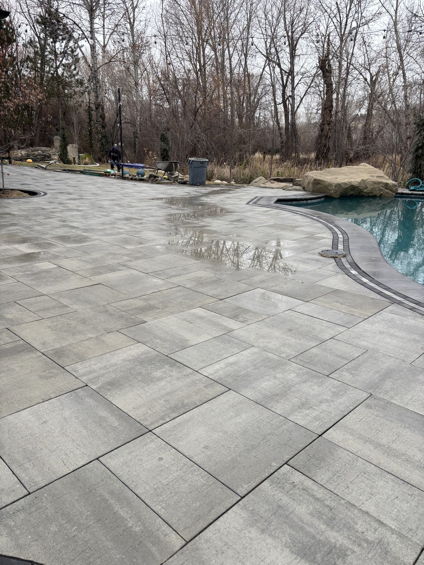A large-format grey paver pool deck curving around the water's edge with a boulder outcrop beyond.
